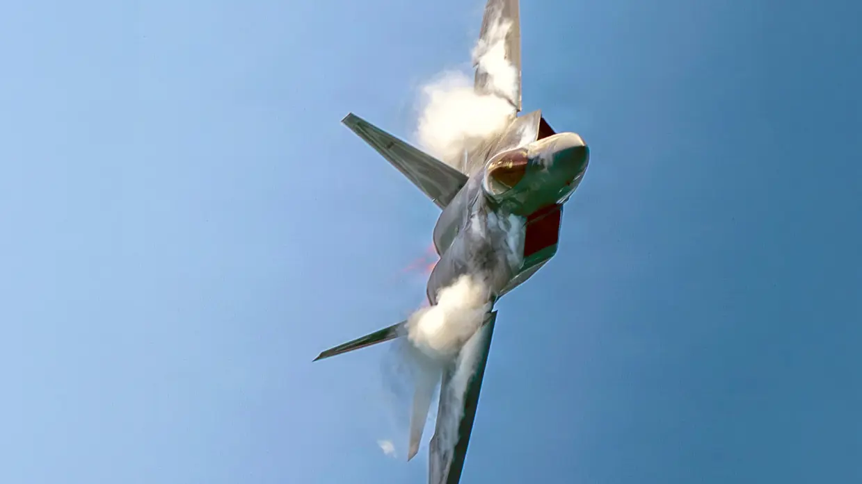 Een F-22 vliegt op zijn kant en maakt een scherpe bocht tegen een blauwe lucht.