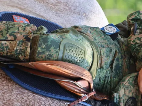 Een soldaat in camouflagekleding met een helm en kniebeschermer zit op een paard met een bruin zadel en teugels.