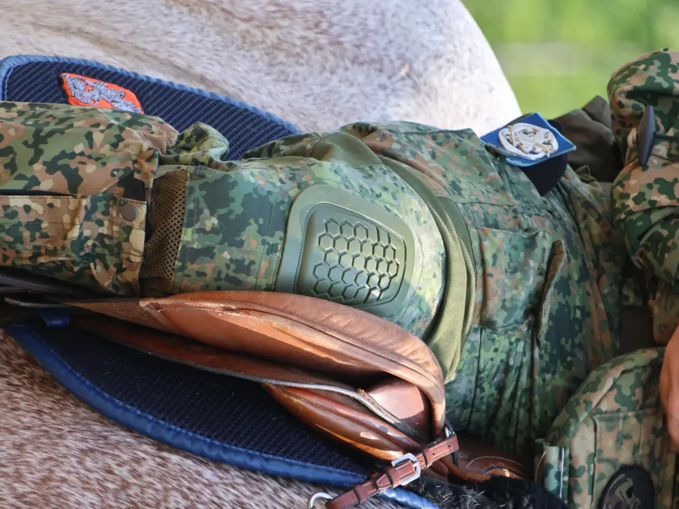 Een soldaat in camouflagekleding met een helm en kniebeschermer zit op een paard met een bruin zadel en teugels.