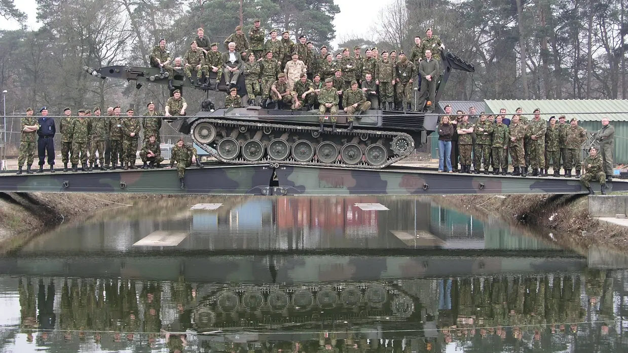 Een grote groep militairen zitten voor de foto op een tank die op een geniebrug staat.