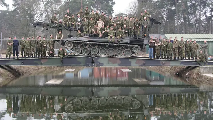 Een grote groep militairen zitten voor de foto op een tank die op een geniebrug staat.