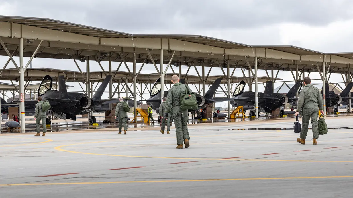 Vliegers lopen met tassen richting F-35’s die onder een overkapping op een platform staan.