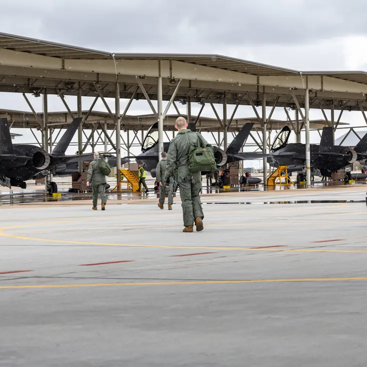 Vliegers lopen met tassen richting F-35’s die onder een overkapping op een platform staan.