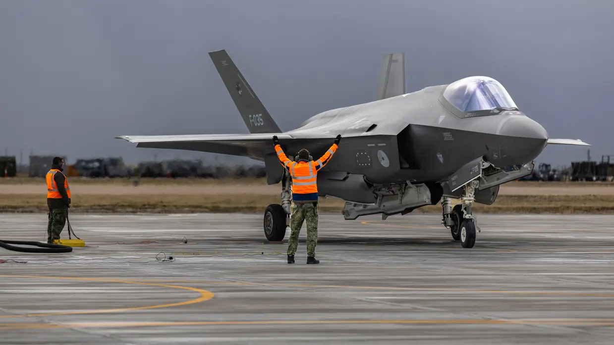 Twee militairen in oranje hesjes naast een F-35 op het platform. Een van de militairen houdt de armen schuin naar boven gespreid in de lucht.