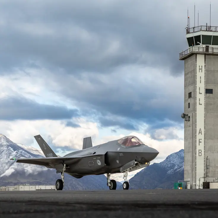 Een F-35 links van de luchtverkeerstoren waarop ‘Hill AFB’ staat. Op de achtergrond besneeuwde bergen.