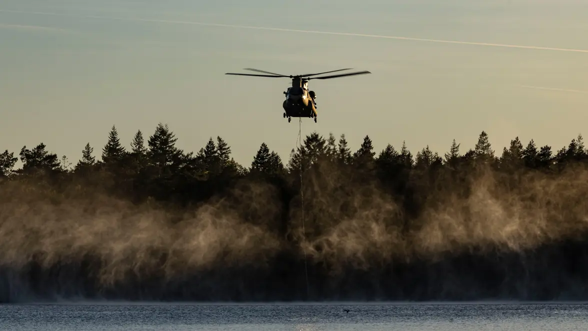 Een Chinook-transporthelikopter hangt laag boven een meer om water te laden in een speciale waterzak.