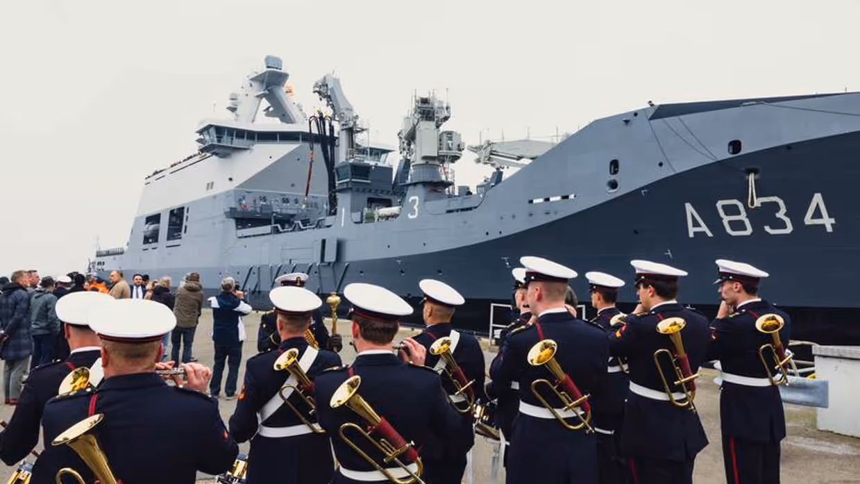 Marineschip ligt langs kade. Op voorgrond militaire muziekkapel.