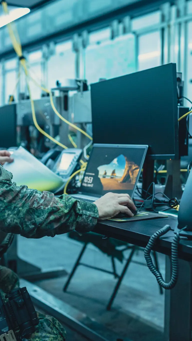 Een bewapende militair zit op een klapstoel achter een computer en bladert door een mapje.
