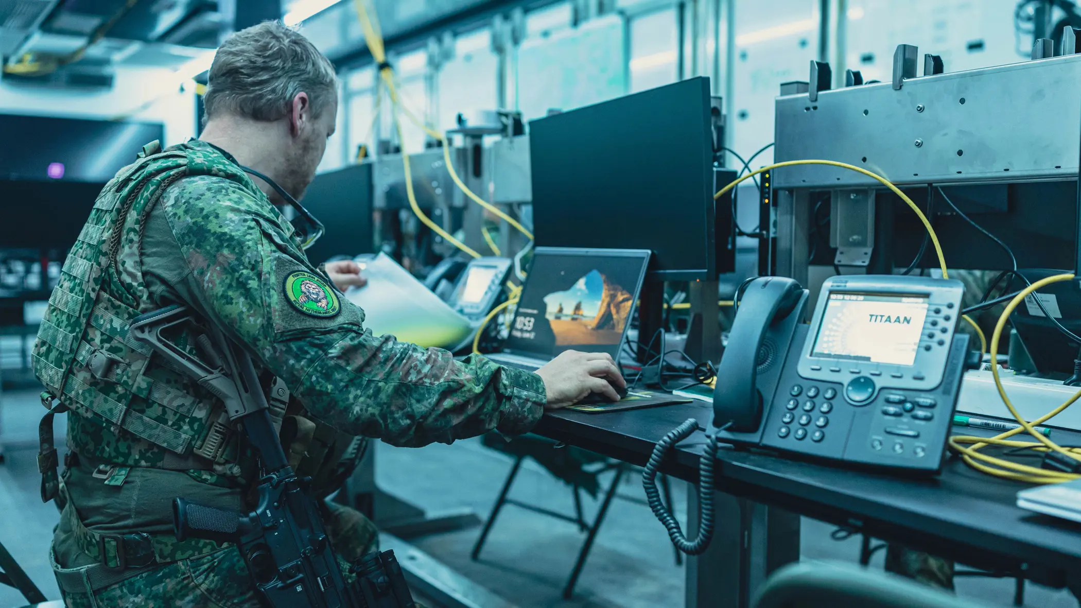 Een bewapende militair zit op een klapstoel achter een computer en bladert door een mapje.