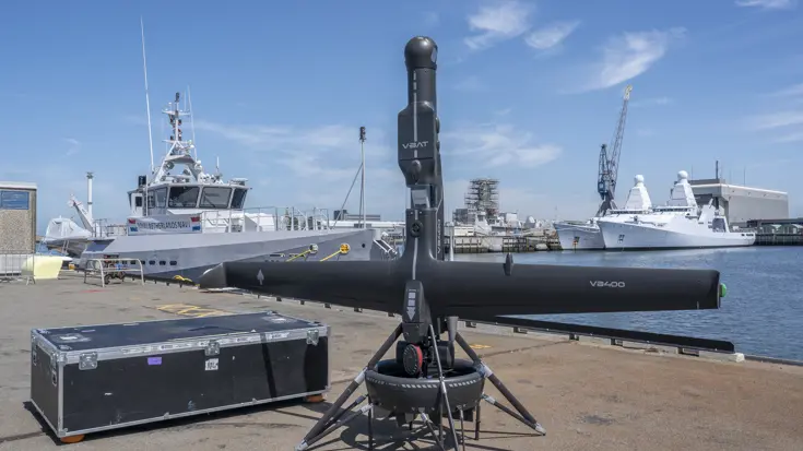 De zwarte V-BAT staat rechtop. Daarachter liggen marineschepen aan de kade.
