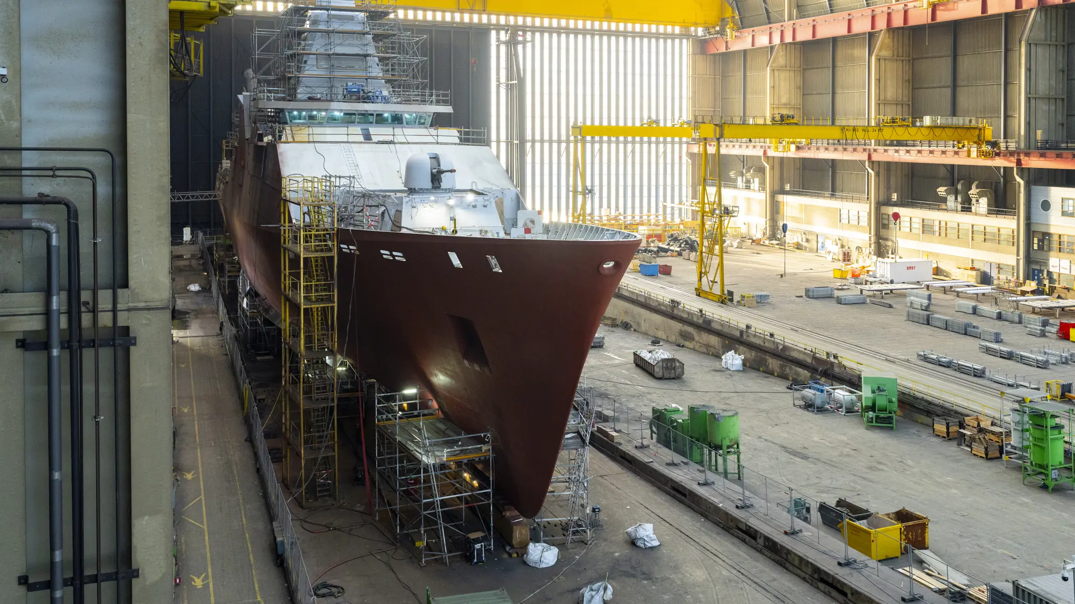 Zr.Ms. Holland in aanbouw in een overdekte scheepswerf, omringd door steigers, kranen en bouwmateriaal. 