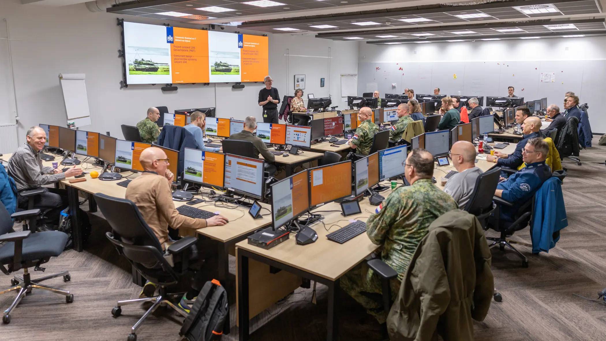 Een ruimte met daarin een dubbele u-opstelling van bureaus. Daaraan zitten militairen en burgers. Zij kijken naar een groot scherm vooraan in de ruimte, waarop een Leopard-gevechtstank te zien is.