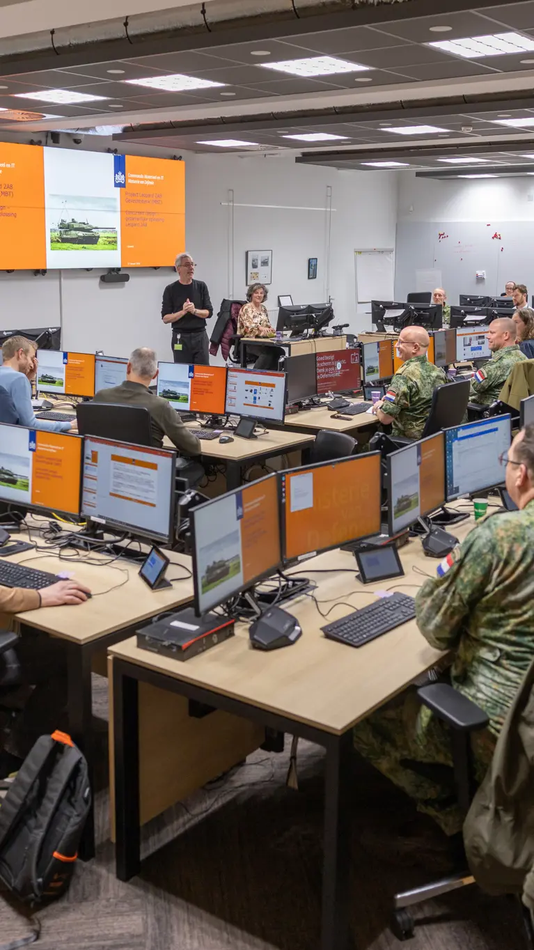 Een ruimte met daarin een dubbele u-opstelling van bureaus. Daaraan zitten militairen en burgers. Zij kijken naar een groot scherm vooraan in de ruimte, waarop een Leopard-gevechtstank te zien is.
