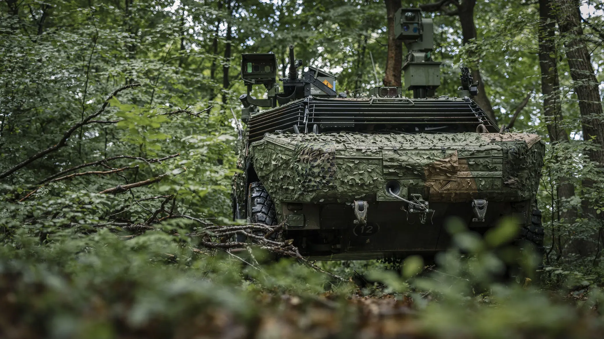 Een Fennek-voertuig rijdt door een beboste omgeving en is voorzien van een camouflagenet. 