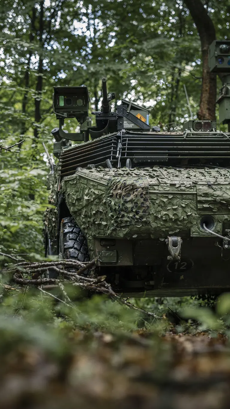 Een Fennek-voertuig rijdt door een beboste omgeving en is voorzien van een camouflagenet. 