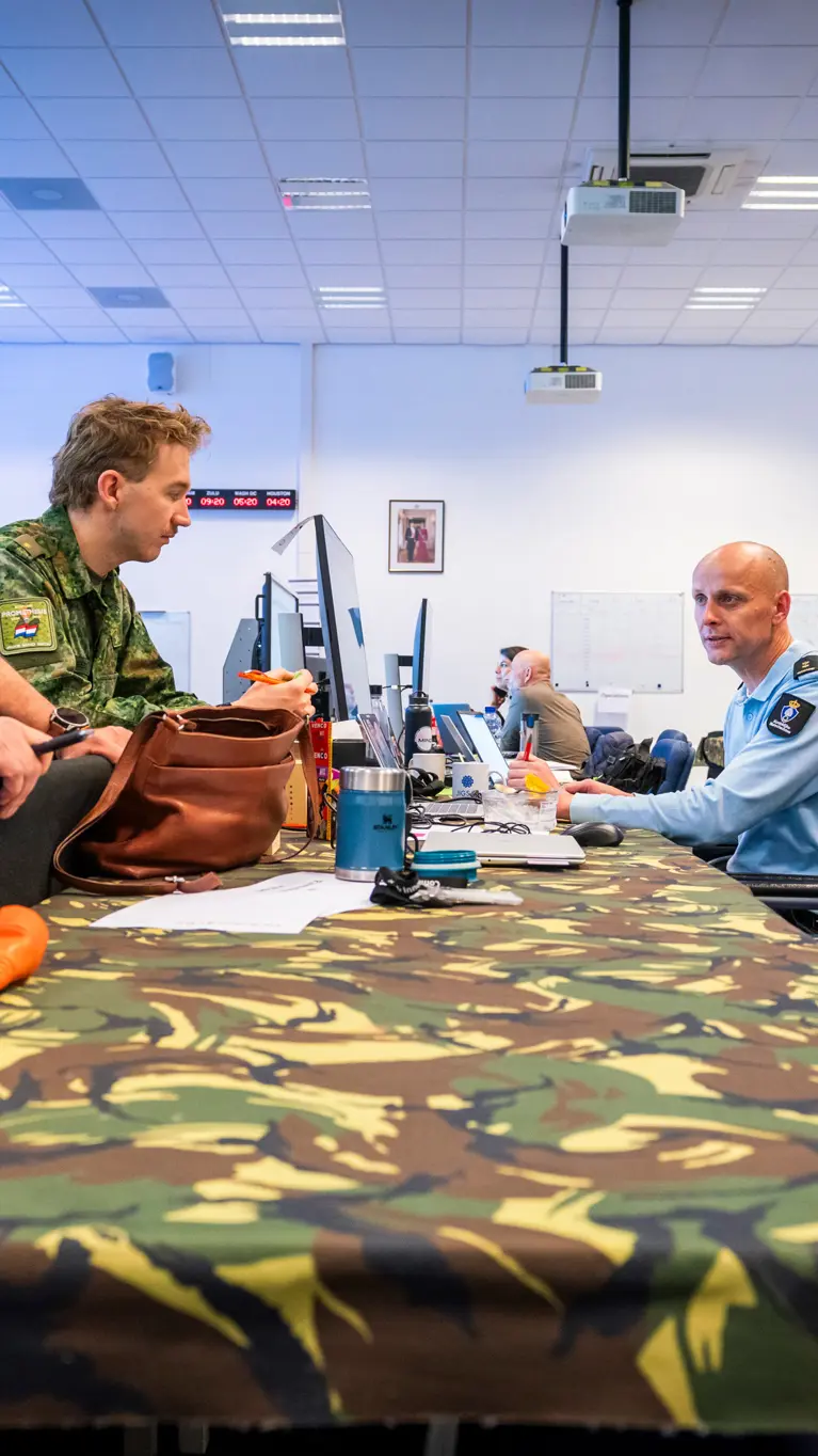Vier militairen en twee burgermedewerkers overleggen staand en zittend aan een gecamoufleerde, bedekte tafel.