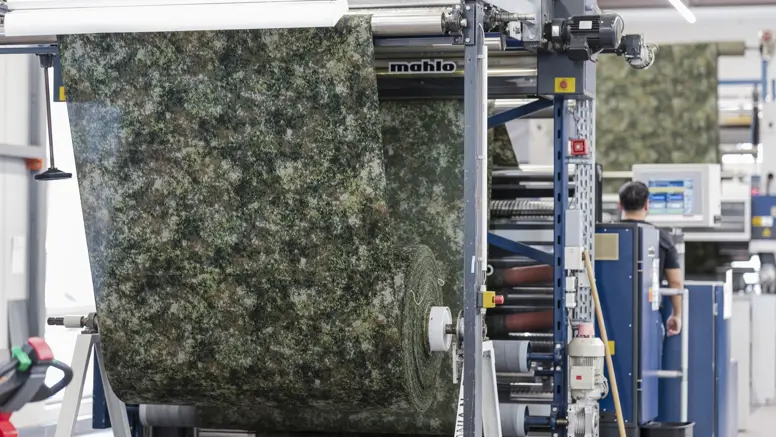 Stof met groen camouflagepatroon wordt door machine geleid tijdens een controle op productiefouten.