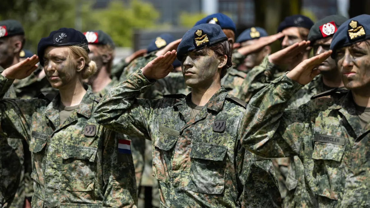 Twee luchtmachtmilitairen brengen samen met militairen van landmacht, marine en marechaussee de militaire groet. Allen met gecamoufleerde gezichten en een baret op. 