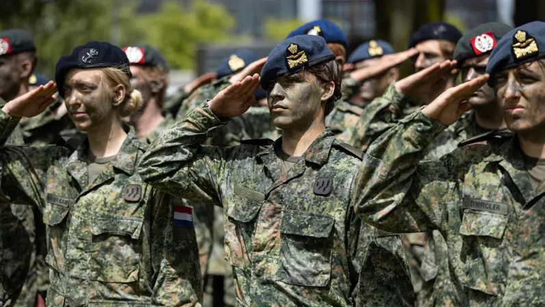 Twee luchtmachtmilitairen brengen samen met militairen van landmacht, marine en marechaussee de militaire groet. Allen met gecamoufleerde gezichten en een baret op. 