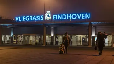De ingang van Vliegbasis Eindhoven, af te lezen op de grote blauwe neonletters boven het gebouw. Daarnaast een vliegende C-390M.