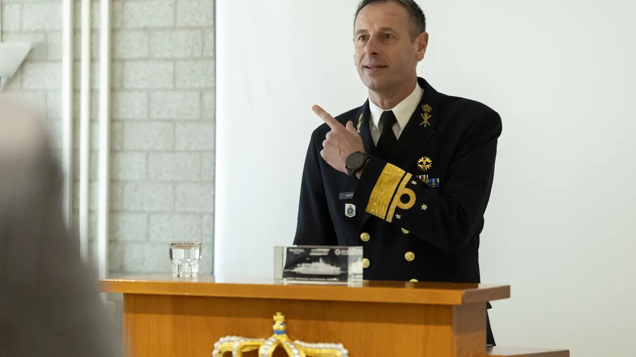 Een sprekende vice-admiraal Jan Willem Hartman, daarnaast zit hij met een burger en een andere militair achter en tafel en zetten zij gelijktijdig een handtekening onder een document.