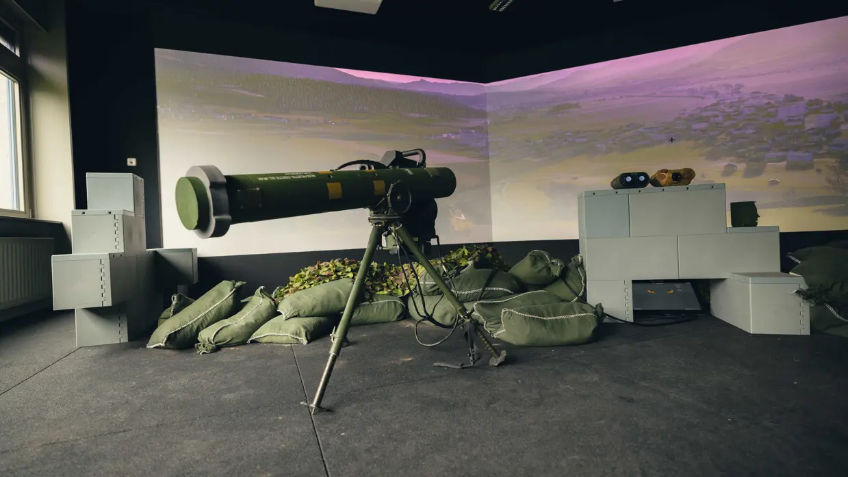Een viertal afbeeldingen. Naast het cilindervormige, donkergroene antitankwapen Spike Long Range gericht op een beamer, hetzelfde middel alleen nu met daarachter een militair. Samen met een instructeur, die achter een beeldscherm zit, bevinden de twee zich in een kleinere ruimte. Vervolgens diezelfde oefenende militair schuin vanaf onder en gezien vanaf voren. Daarbij is zijn geconcentreerde blik zichtbaar.