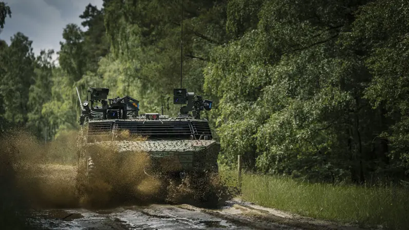 Een militair voertuig raast in het bos over een zandweg. 