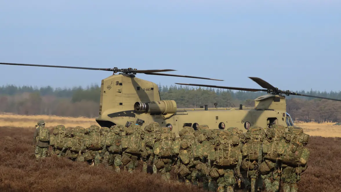 Een grote groep militairen met rugzakken en wapens lopen naar de achterzijde van een grote helikopter op de heide.