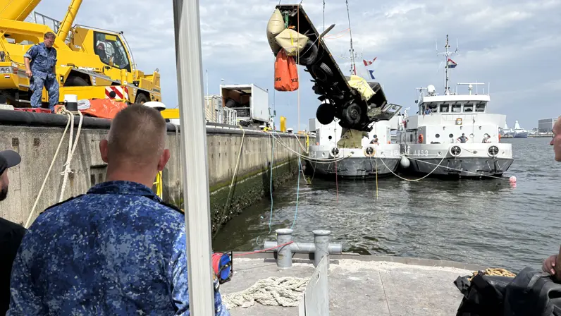 Een wrak wordt met een takel geborgen uit het water. Voor de cameraman staat iemand met een blauwgevlekt marinepak.