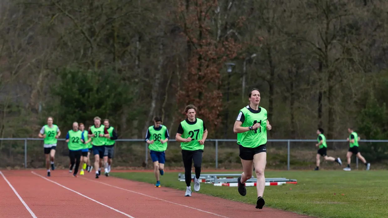 Een groep hardlopers in gele hesjes met nummers rent op een atletiekbaan. De voorste loper lijkt gefocust, met bomen en een hek op de achtergrond.