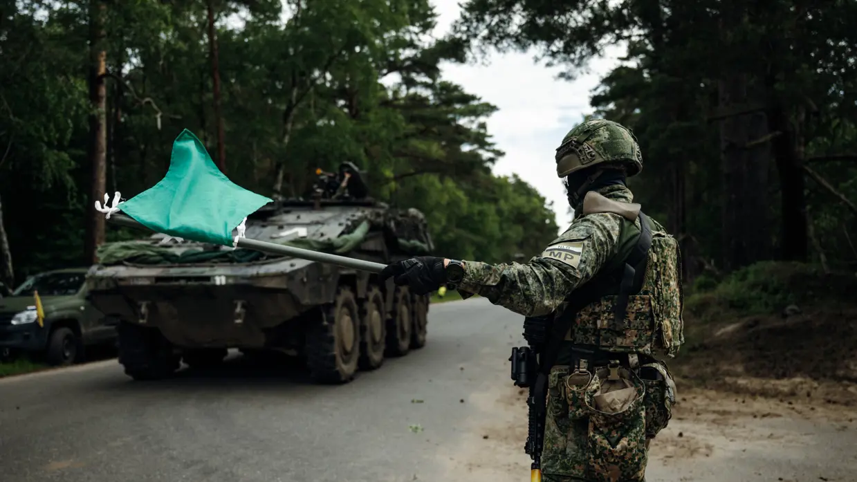 Marechaussee in groen tenue met een groene vlag in zijn hand, op de achtergrond een voertuig van de Landmacht.