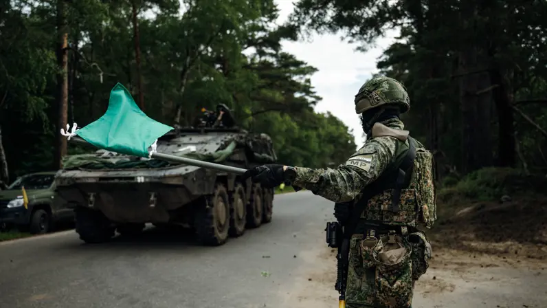 Marechaussee in groen tenue met een groene vlag in zijn hand, op de achtergrond een voertuig van de Landmacht.