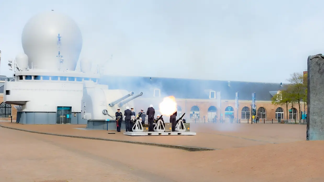 Een wit schip met radarbol, kanonnen en uniformen: een saluutschot klinkt op een open plein met bomen. 