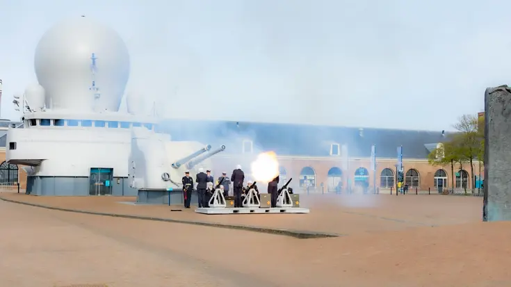 Een wit schip met radarbol, kanonnen en uniformen: een saluutschot klinkt op een open plein met bomen. 