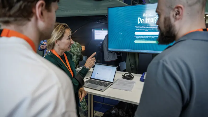 Drie personen voeren een gesprek voor een laptopscherm en een groot beeldscherm.