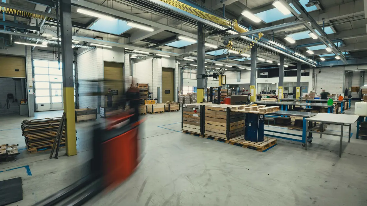 Een grijze loods met daarin diverse lange tafels en een langsrijdende heftruck.