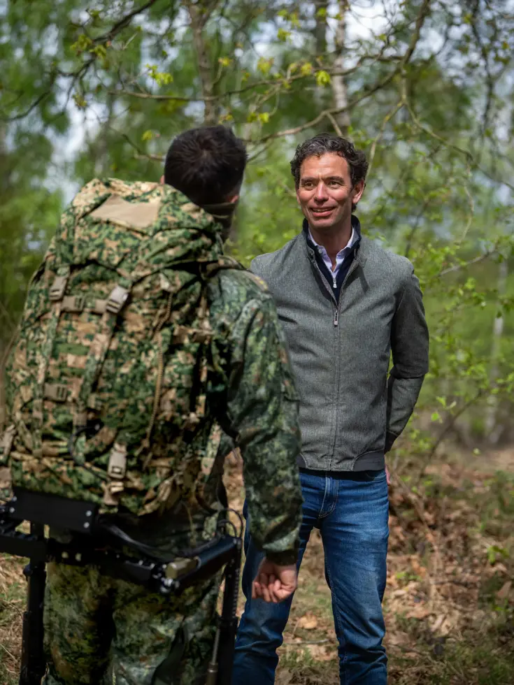 Een man in grijs jack praat met een soldaat in camouflage en rugzak in een groen bos. 
