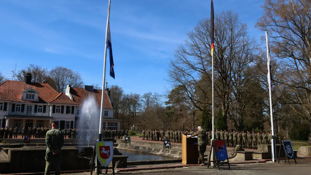 Een militair staat achter een spreekgestoelte met de Nederlands en Duitse vlag in top, met veel militairen in gelid op de achtergrond.