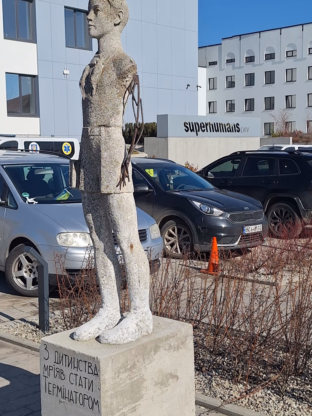 Een groot, liggend stenen beeld van een persoon, half ingebed in een betonnen blok, naast een modern gebouw met geparkeerde auto’s en een heldere lucht.