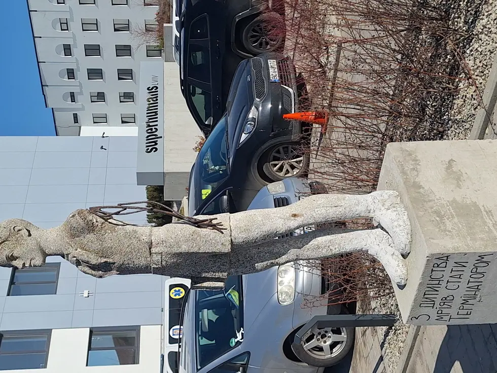 Een groot, liggend stenen beeld van een persoon, half ingebed in een betonnen blok, naast een modern gebouw met geparkeerde auto’s en een heldere lucht.