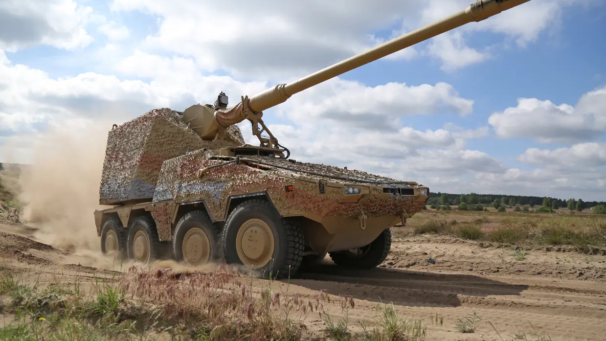Een zwaar militair pantservoertuig met een lang kanon rijdt snel over een zanderig terrein, terwijl stof en zand opwaaien onder een blauwe, bewolkte hemel. 