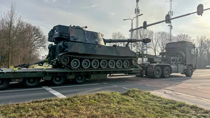 Een groene pantserhouwitser staat op een lage oplegger, getrokken door een zware vrachtwagen op een weg met verkeerslichten. 