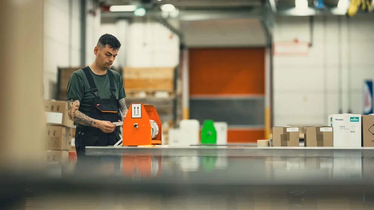 Een man aan een lange tafel is bezig met stickers. Op de tafel liggen diverse dozen.&nbsp;