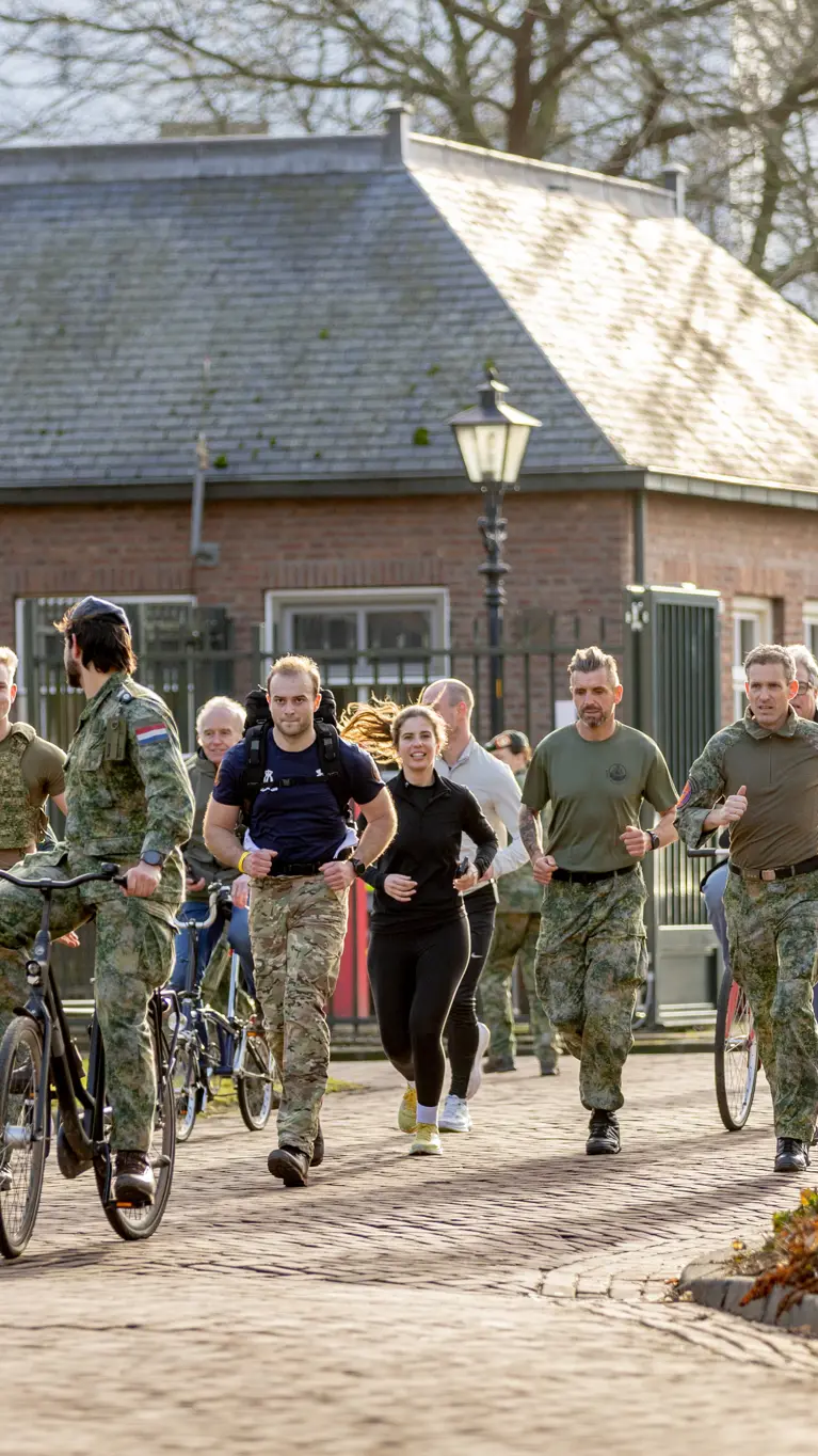 Een groep mensen in beweging op een zonnig pad: sommigen lopen, anderen fietsen. Ze dragen sportieve kleding en militaire uniformen, met bomen en een gebouw op de achtergrond.