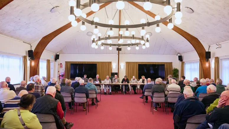 In een oude zaal zitten oudere mensen en achter de tafel een man met een hamer en serieus kijkende mensen.