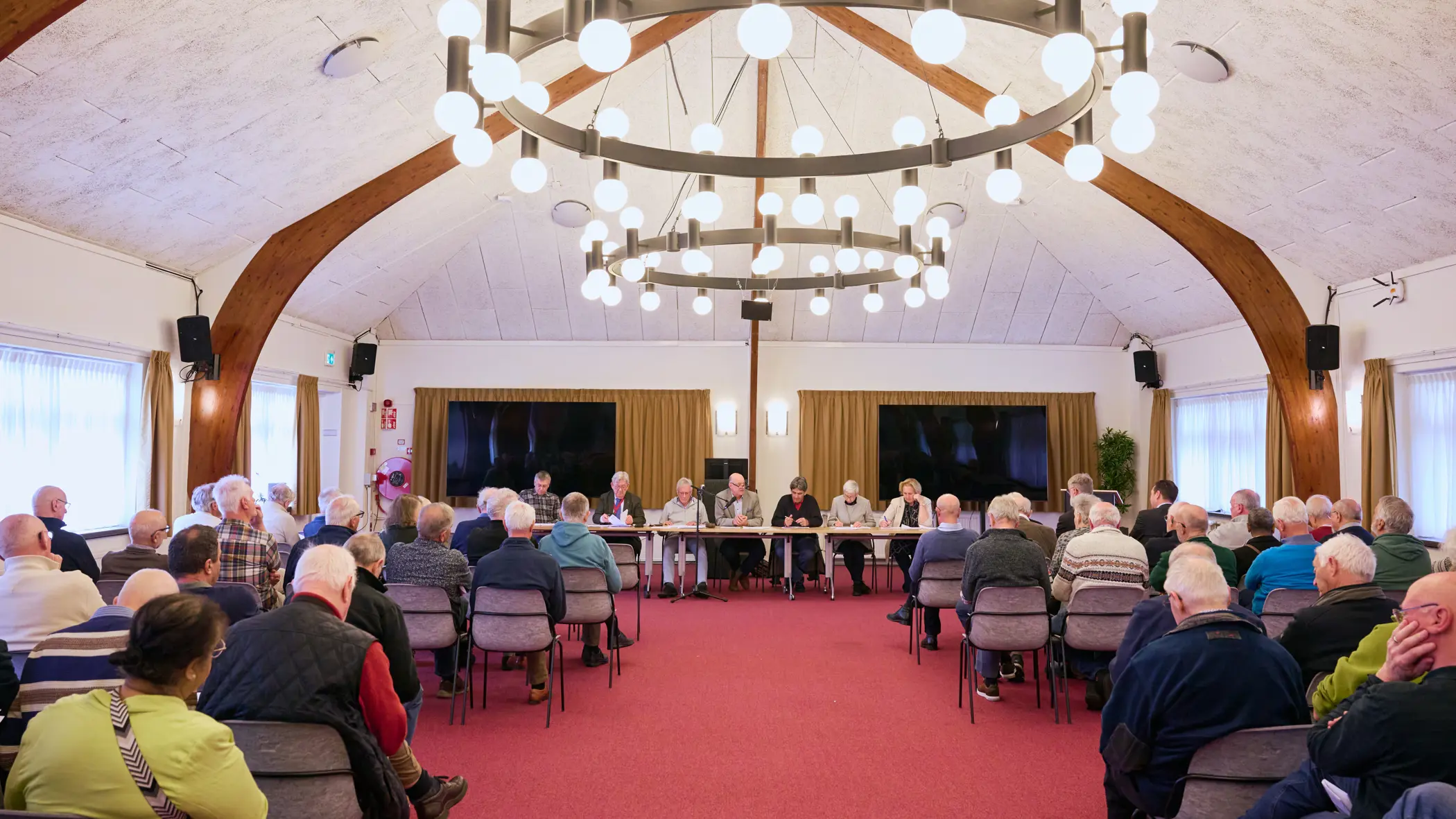In een oude zaal zitten oudere mensen en achter de tafel een man met een hamer en serieus kijkende mensen.