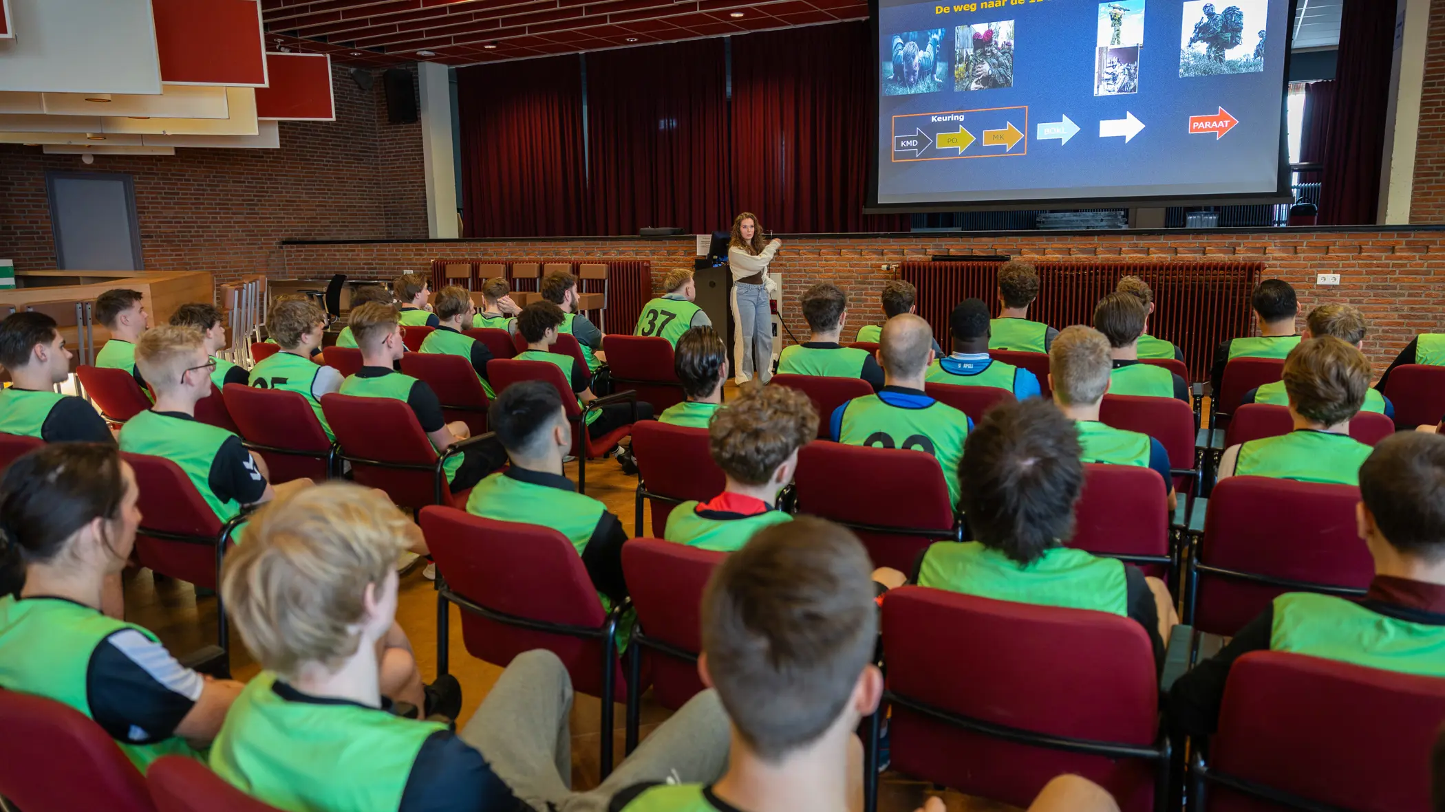 In een zaal houdt een vrouw een voordracht over de luchtmobiele brigade. Studenten in gele vesten luisteren aandachtig naar de presentatie op een groot scherm.&nbsp;