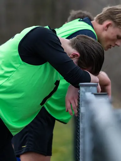 Een groep hardlopers in gele hesjes met nummers rent op een atletiekbaan. De voorste loper lijkt gefocust, met bomen en een hek op de achtergrond.