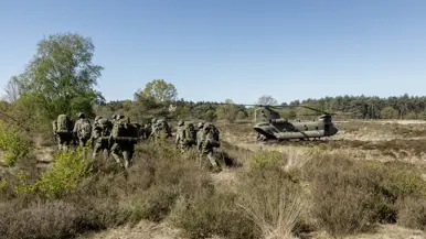 Een groep soldaten in camouflage loopt door een heideveld met lage struiken, een helikopter staat op de achtergrond. 