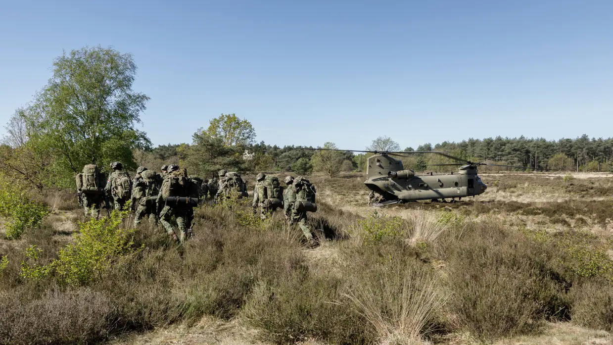 Een groep soldaten in camouflage loopt door een heideveld met lage struiken, een helikopter staat op de achtergrond. 